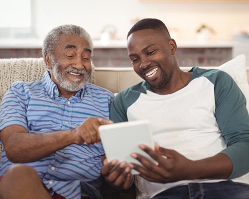 a grown child and their parent sitting on the couch and looking at an electronic tablet